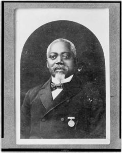 Black and white photo; African American man with white hair and goatee looks head on at camera. Wearing suit with medals.